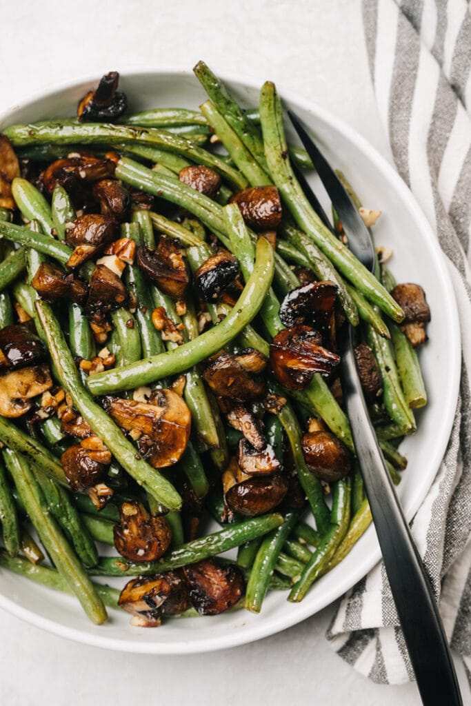 Roasted green beans and mushrooms in a white serving bowl with a silver serving fork and striped linen napkin to the side.