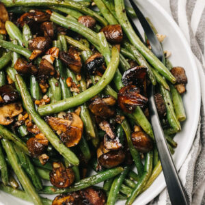 Roasted green beans and mushrooms in a white serving bowl with a silver serving fork and striped linen napkin to the side.
