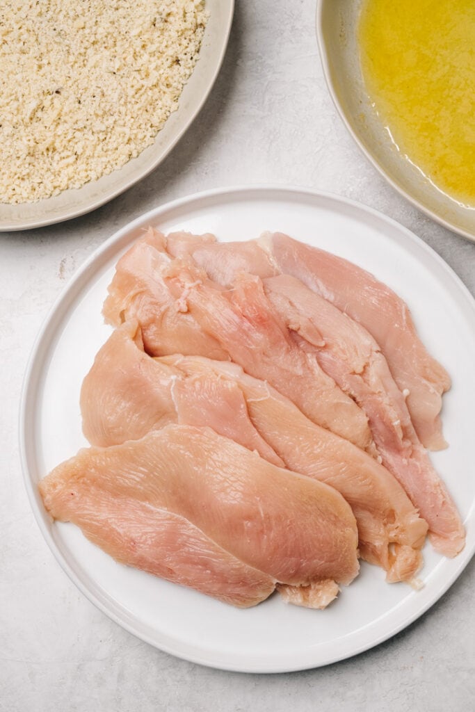 Thinly sliced chicken breasts on a white platter with a bowl of melted butter and a bowl of seasoned breadcrumbs to the side.