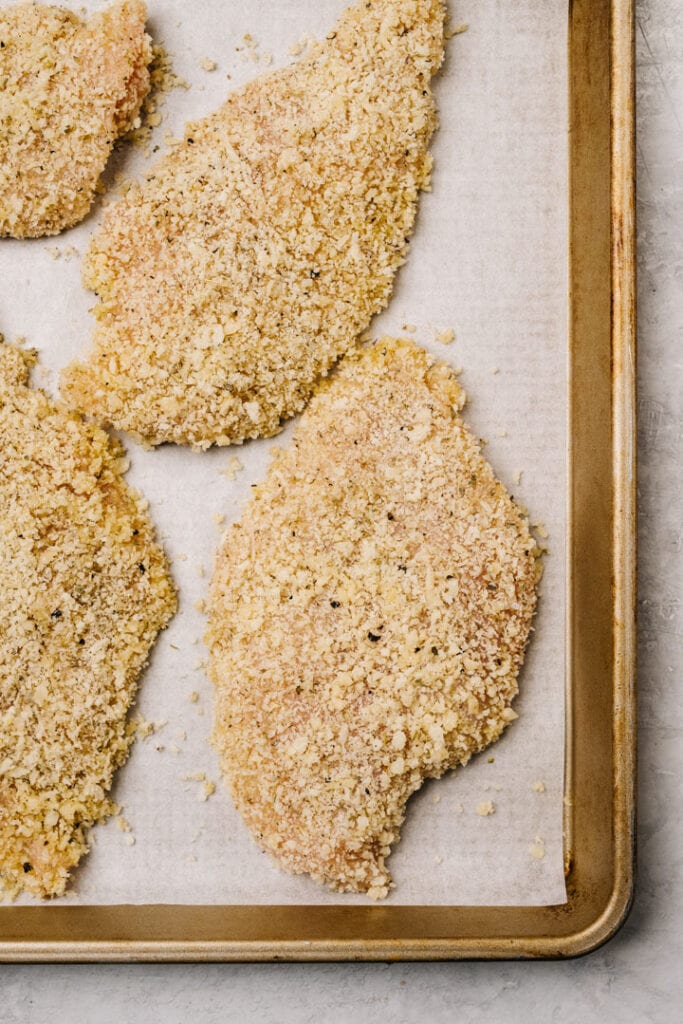 Thinly sliced chicken breasts coated with parmesan on a parchment lined baking sheet.