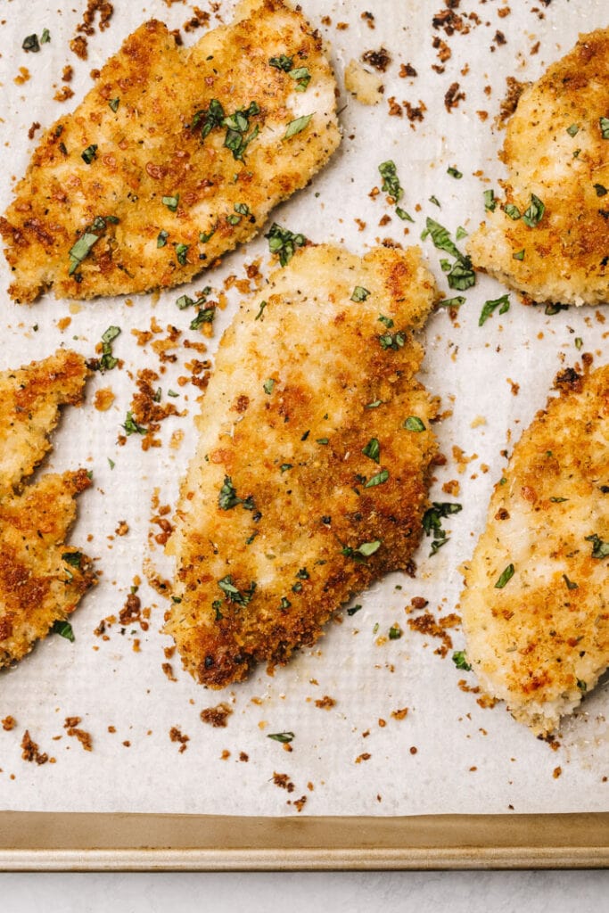 Golden brown parmesan crusted chicken breasts on a parchment lined baking sheet, garnished with chopped parsley.