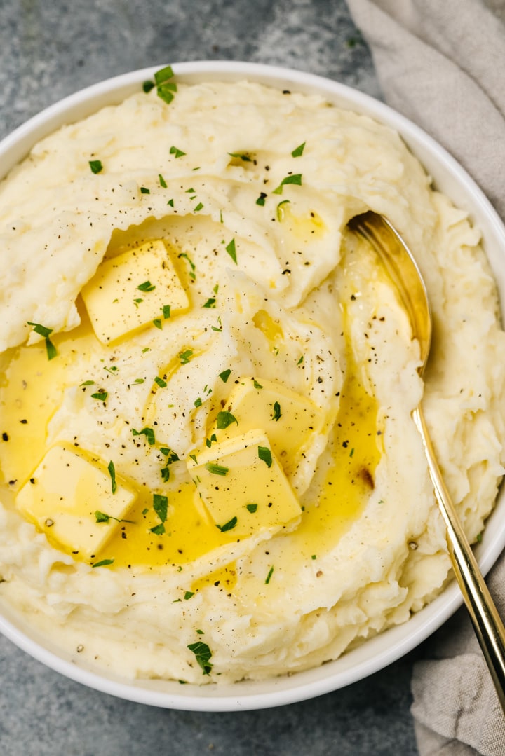 A gold serving spoon tucked into a bowl of fluffy roasted garlic mashed potatoes, topped with melting butter and fresh herbs.
