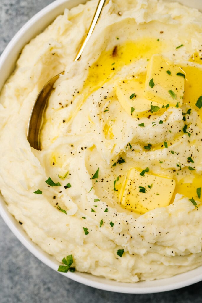 Roasted garlic mashed potatoes in a serving bowl with a gold spoon, drizzled with butter and chopped fresh herbs.