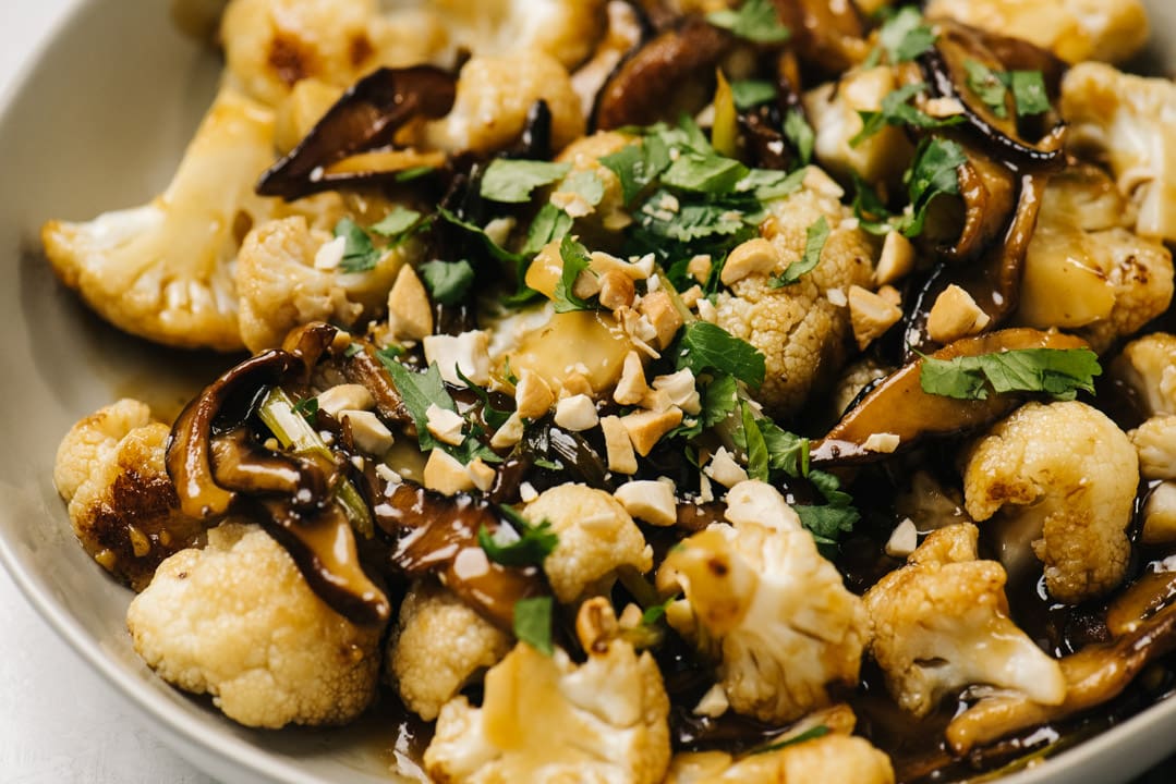 Side view, cauliflower stir fry with mushrooms in a low tan serving bowl, garnished with chopped cashews and fresh cilantro.