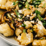 Side view, cauliflower stir fry with shiitake mushrooms in a tan serving dish, garnished with cilantro and chopped cashews.