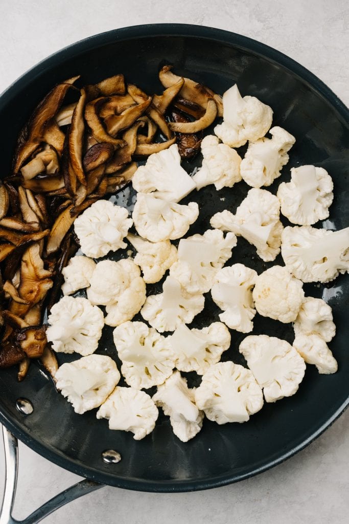 Bite sized cauliflower florets added to shiitake mushrooms in a large skillet.