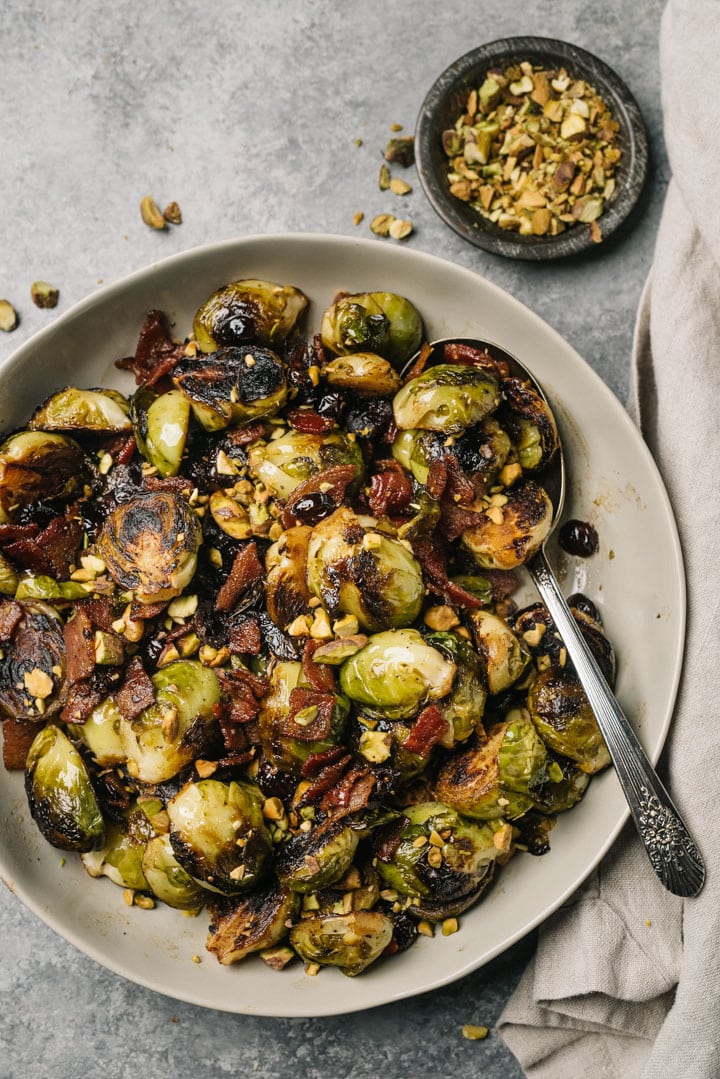 A large tan serving bowl of Christmas themed sauteed brussels sprouts with bacon, cranberries, and pistachios.
