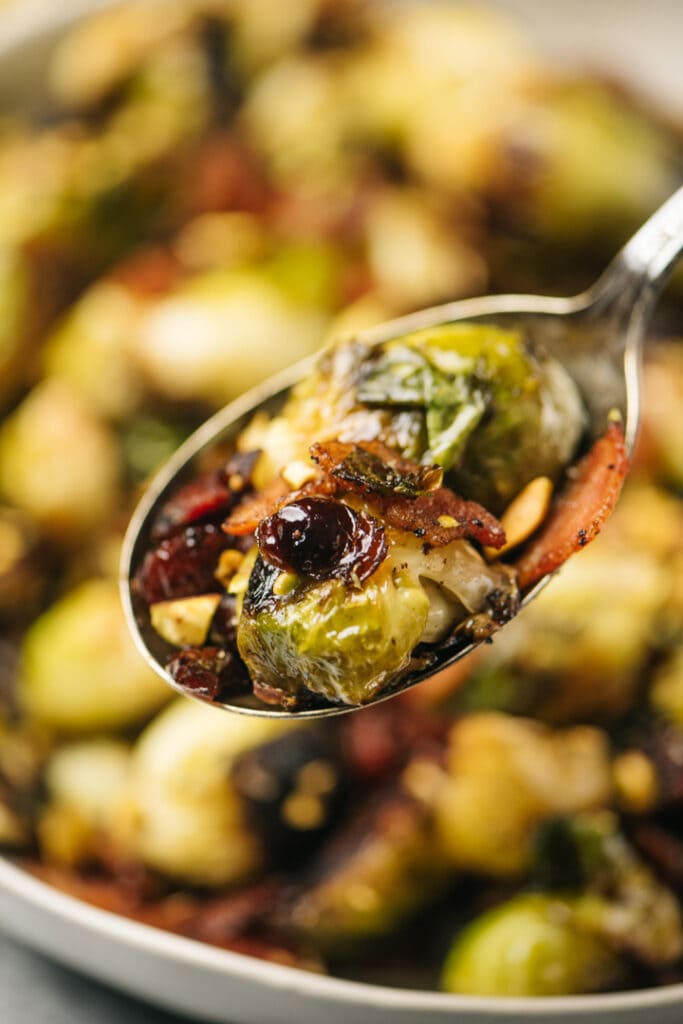 Sauteed brussels sprouts with bacon, cranberries and pistachios on a serving spoon hovering over a serving bowl.