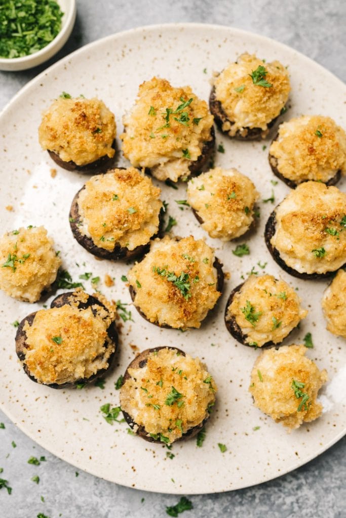 Stuffed mushrooms with crab and cream cheese arranged on a speckled cream serving platter.