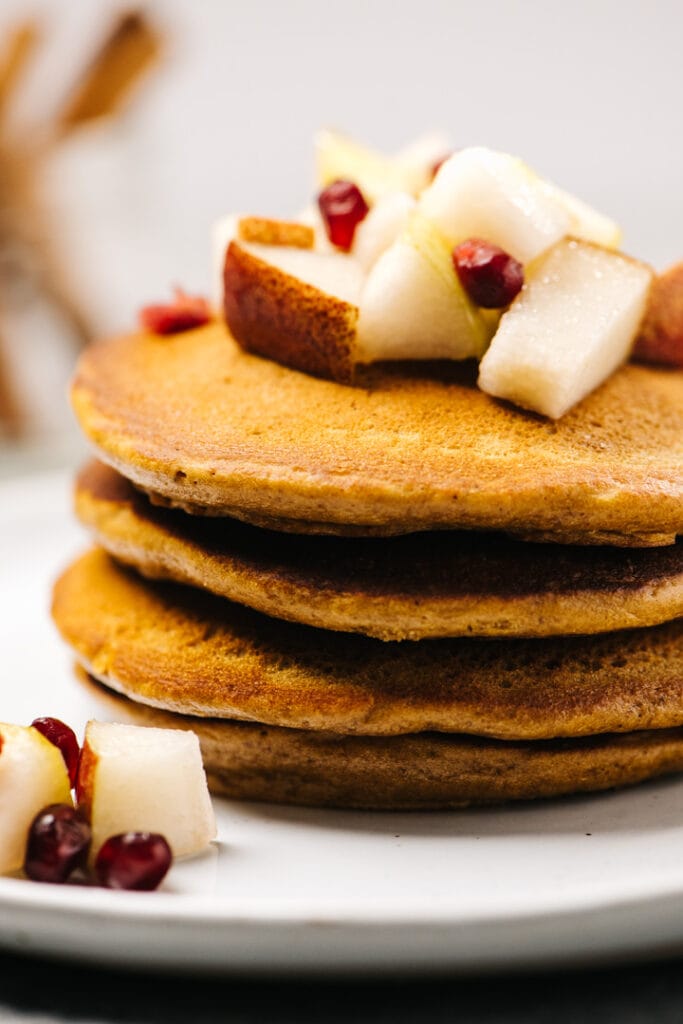 Side view, gingerbread pancakes on a white plate topped with diced pears and pomegranate seeds.