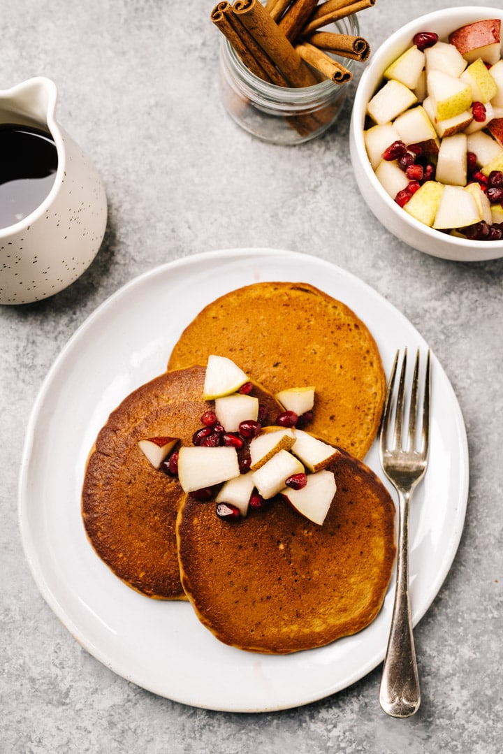 Three gingerbread pancakes on a white plate topped with fresh cut pears and pomegranate seeds.