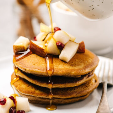 Pouring maple syrup onto a stack of gingerbread pancakes toped with diced pears and pomegranate seeds.
