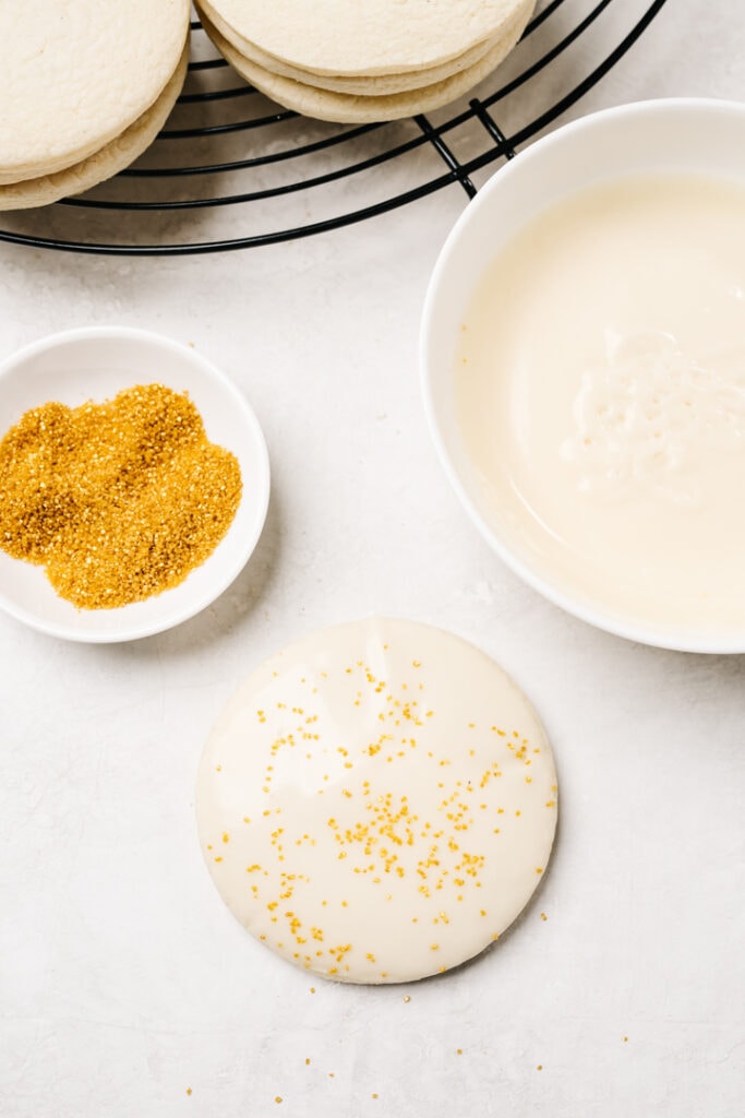 An iced sugar cookie with gold sprinkles surrounded by more sugar cookies, a bowl of vanilla glaze, and a small bowl of gold sprinkles.