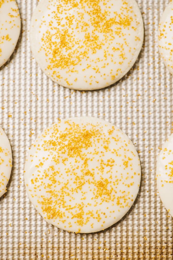 Gluten free sugar cookies with vanilla glaze and gold sprinkles on a rose-gold baking sheet.