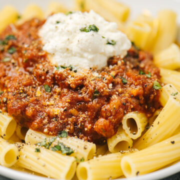 Homemade meat sauce over rigatoni pasta in a shallow white pasta bowl, garnished with ricotta cheese, chopped basil, and ground pepper.