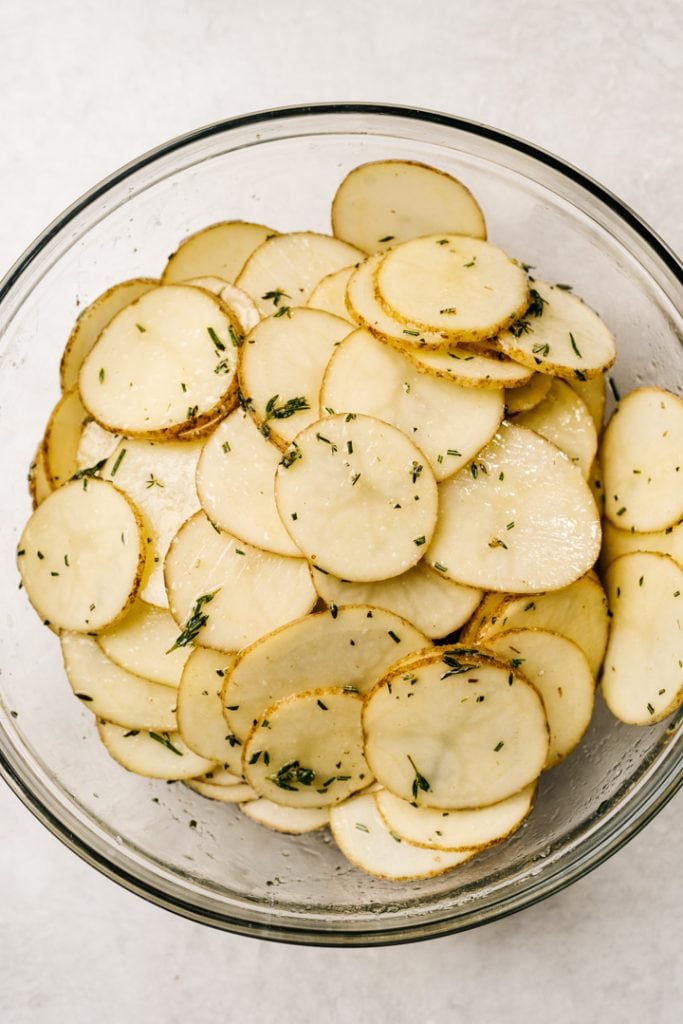 Thinly sliced potatoes tossed with melted butter, olive oil, and fresh minced herbs in a glass mixing bowl.