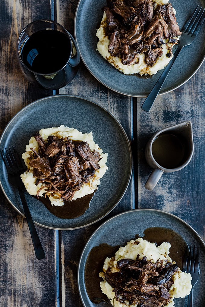 Three servings of pot roast with red wine over mashed potatoes on a wood table, with a small pitch of au just and several glasses of red wine.
