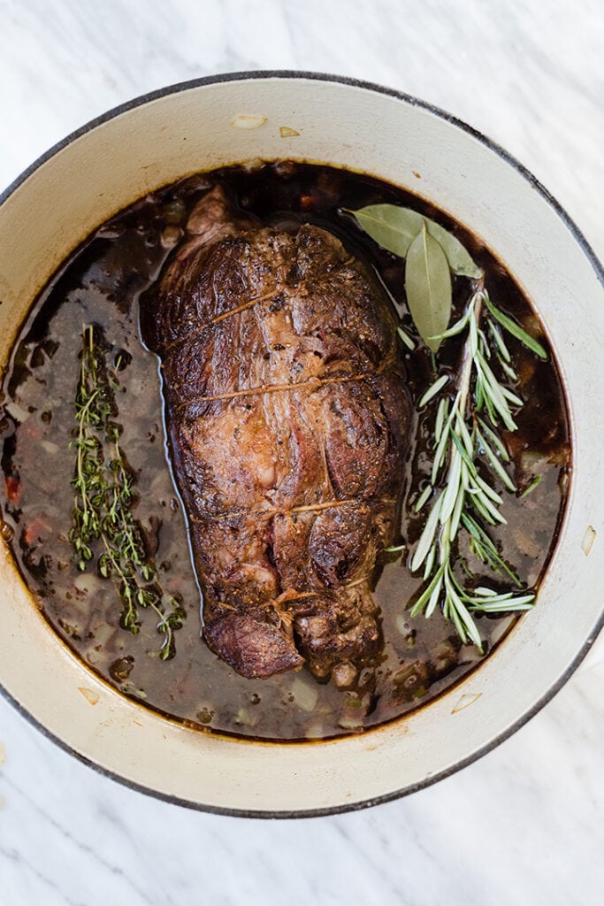 A browned chuck roast in a white dutch oven with fresh herbs and red wine braising liquid.