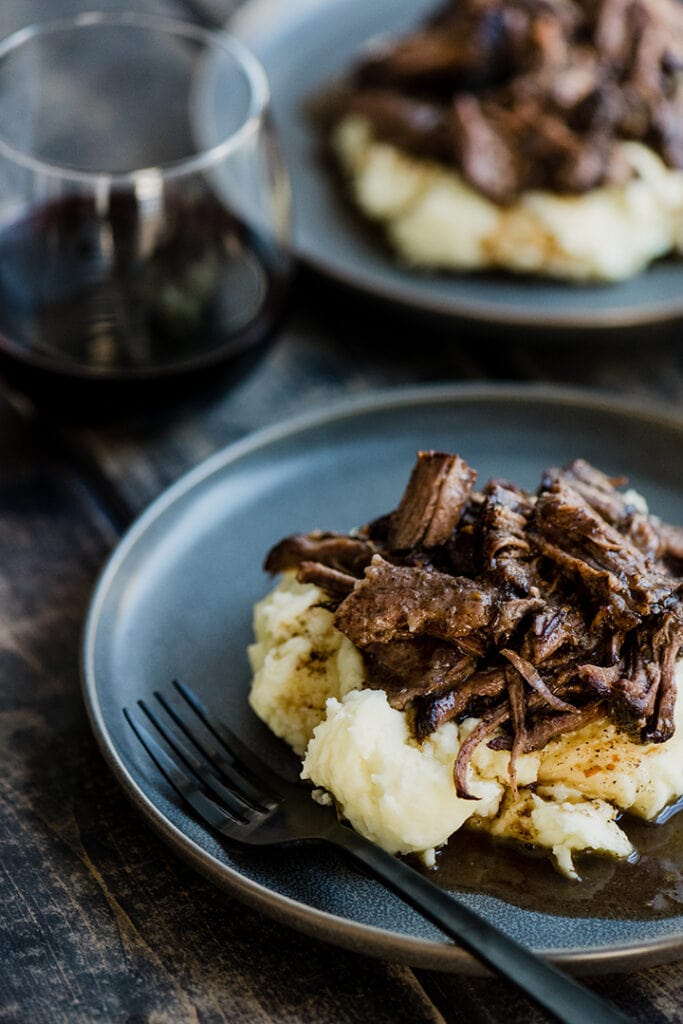 Side view, two plates of pot roast with red wine over mashed potatoes on a wood table with a glass of red wine.