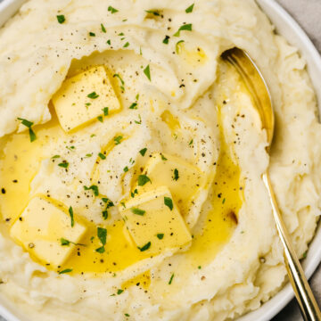 Roasted garlic mashed potatoes in a serving bowl with a gold serving spoon, garnished with parsley and melting butter.