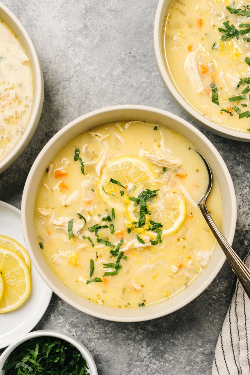 Several bowls of low carb greek chicken soup (avgolemono) on a concrete background, garnished with lemon slices and chopped parsley.