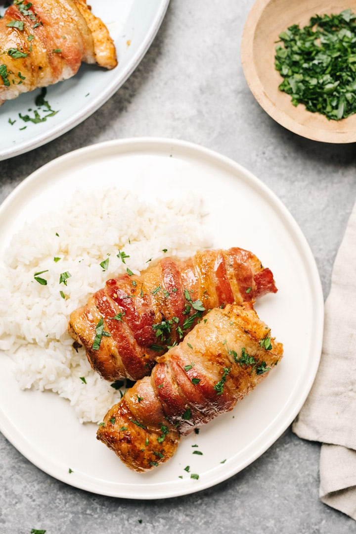 Two pieces of bacon wrapped chicken over white rice on a plate, with a linen napkin and small bowl of chopped parsley to the side.