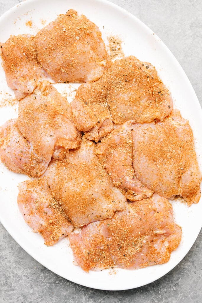 Raw chicken thighs coated in dry rub on a white platter.