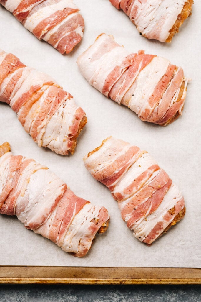 Seasoned chicken thighs wrapped with bacon on a parchment lined baking sheet.