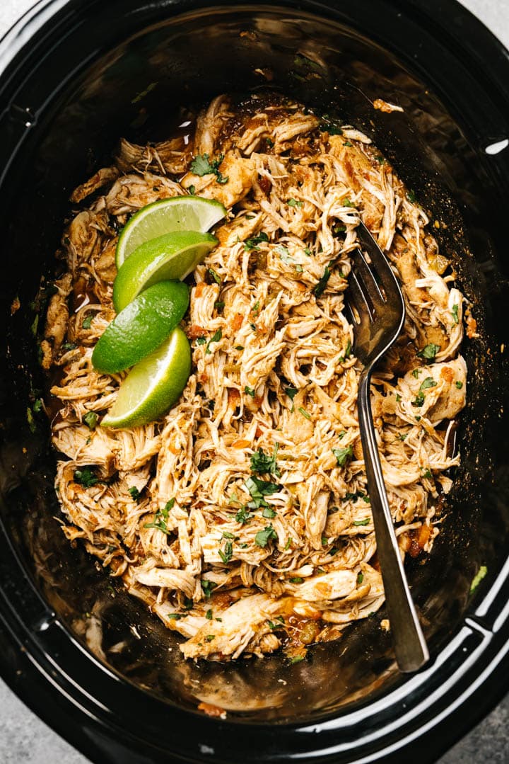 Shredded salsa chicken in a crockpot garnished with cilantro and lime wedges with a serving fork tucked into the chicken.