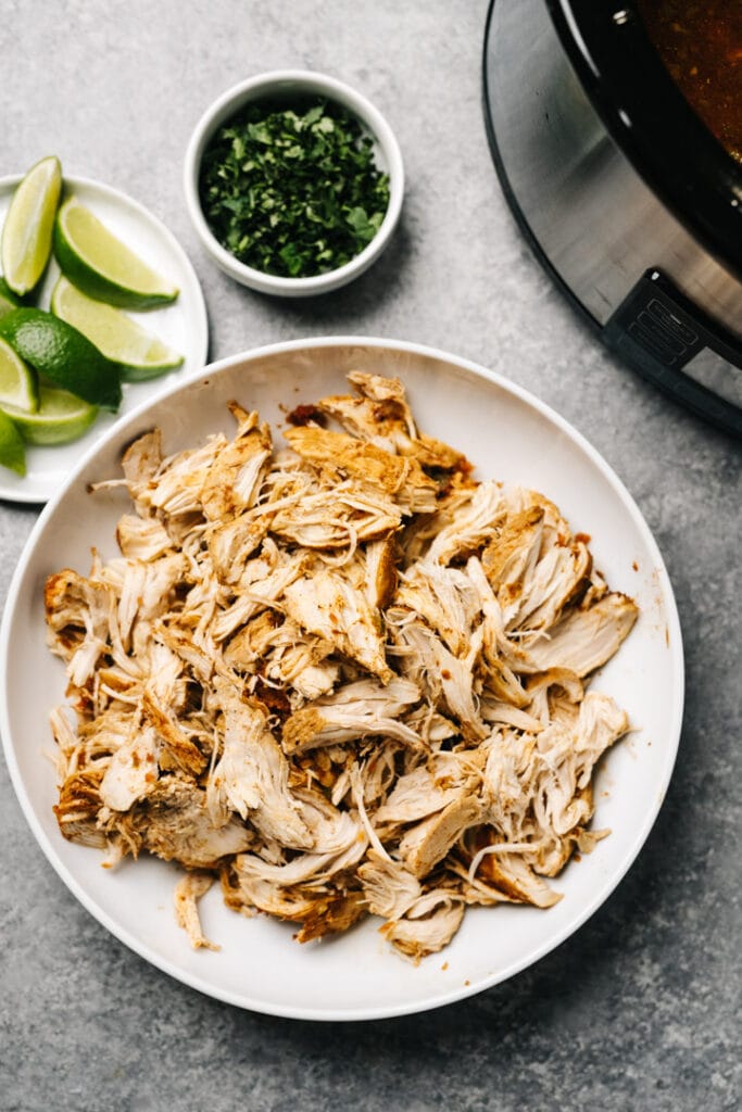 Shredded salsa chicken in a white bowl, with lime wedges, chopped cilantro, and a slow cooker in the background.