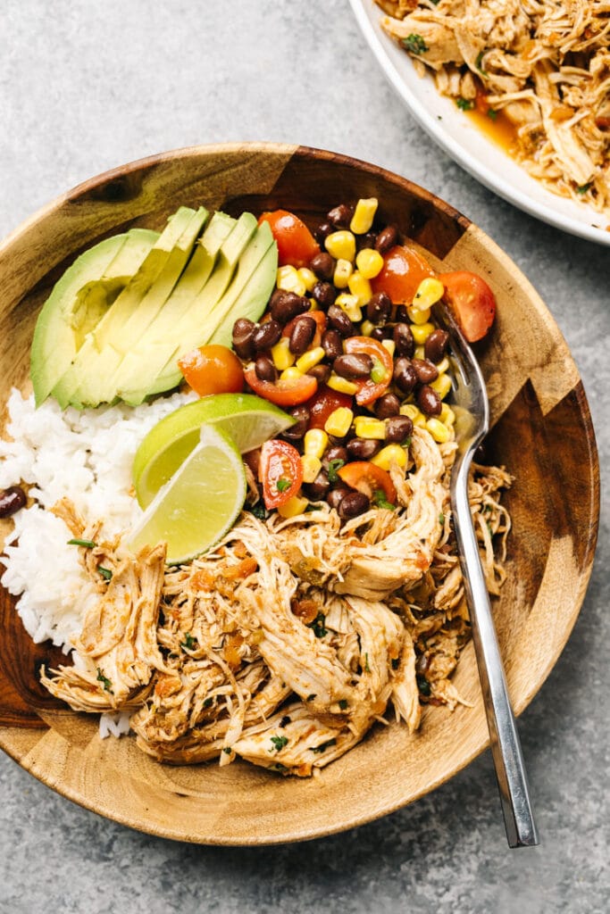 Slow cooker salsa chicken in a wood bowl over rice with back bean salsa, avocado slices, and lime wedges.