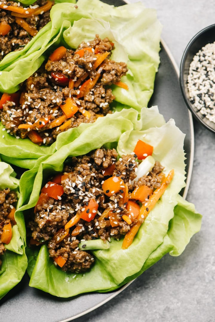 Korean style ground beef lettuce wraps on a black plate with a small bowl of sesame seeds to the side.