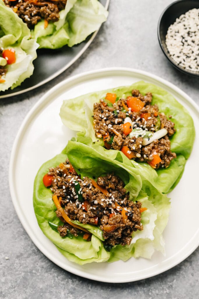 Two asian lettuce wraps with ground beef on a white plate, with a platter of wraps and small bowl of sesame seeds in the background.