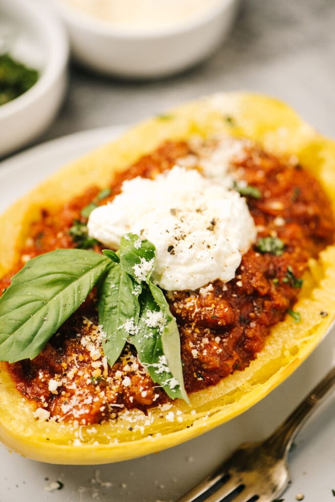 Side view, meat sauce in a roasted spaghetti squash bowl, topped with ricotta cheese, basil, and grated parmesan cheese.