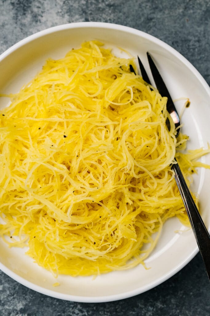 Strands of roasted spaghetti squash in a low white bowl with a silver serving fork.