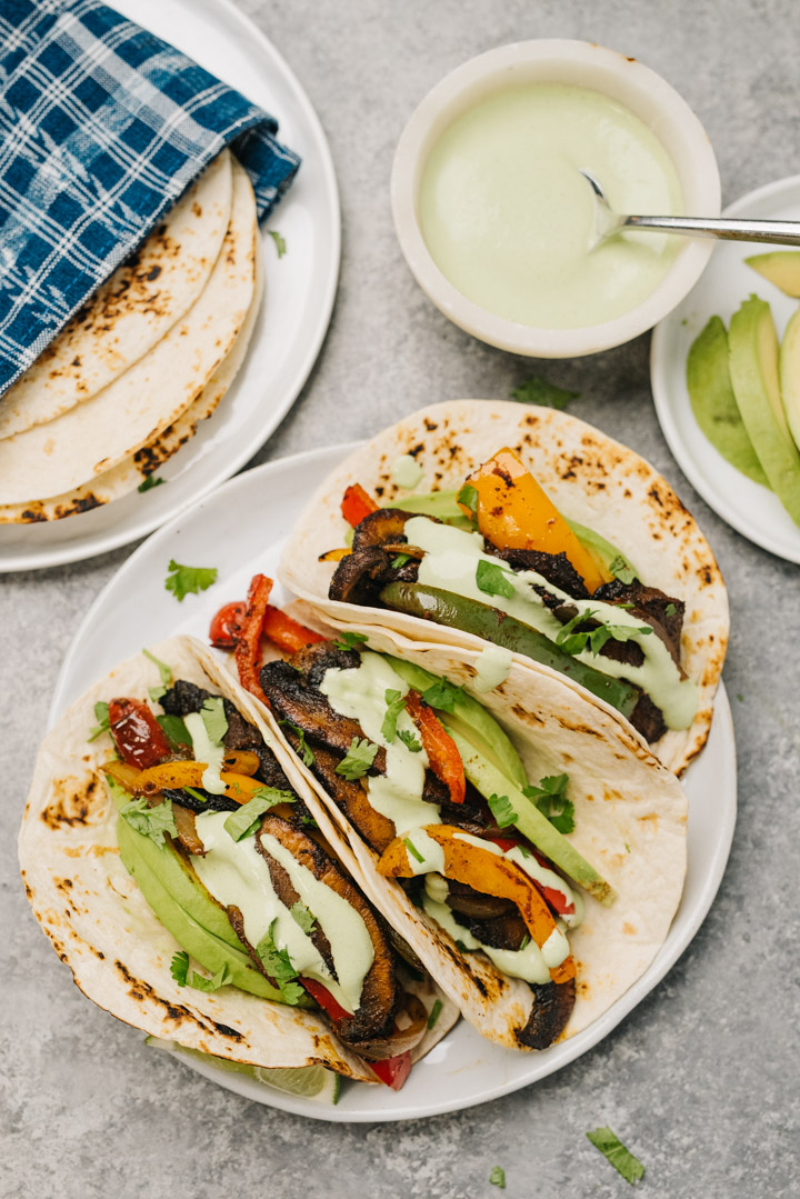 Three vegetarian fajitas on a white plate, stuffed with portobello mushrooms, onions, peppers, and avocado slices, garnished with lime wedges and cilantro lime crema.