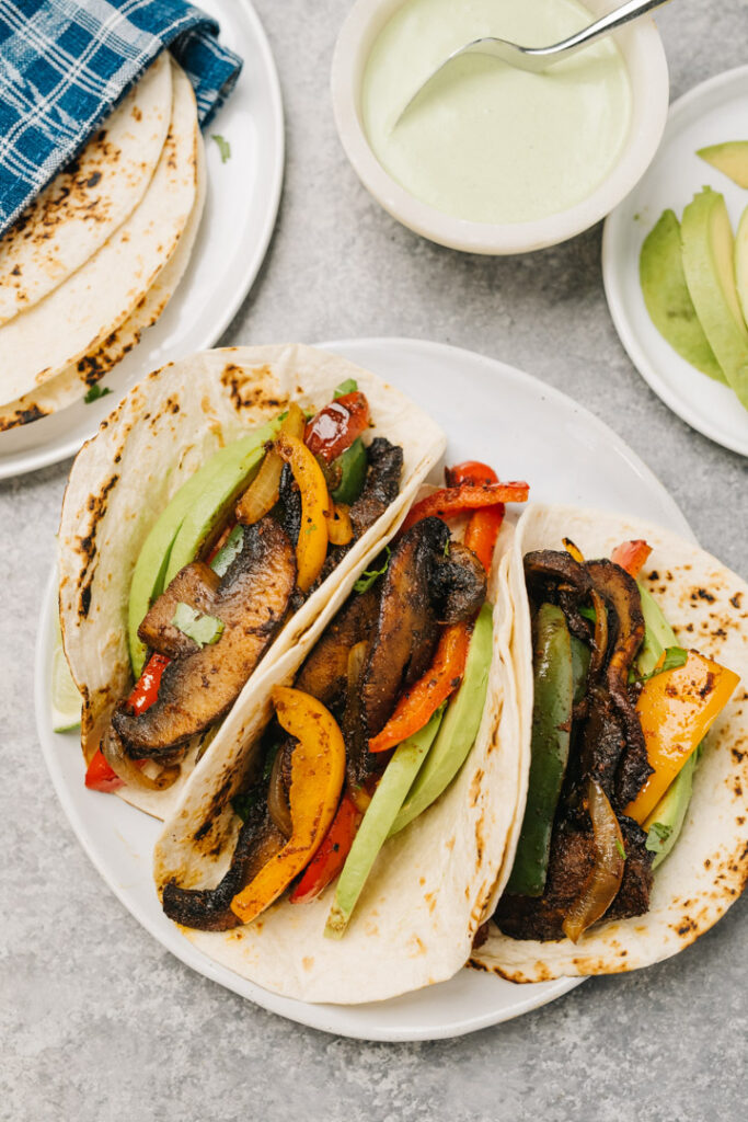 Three vegetarian fajitas filled with mushrooms, peppers, onions, avocado slices and limes, with a bowl of cilantro sour cream and flour tortillas to the side.