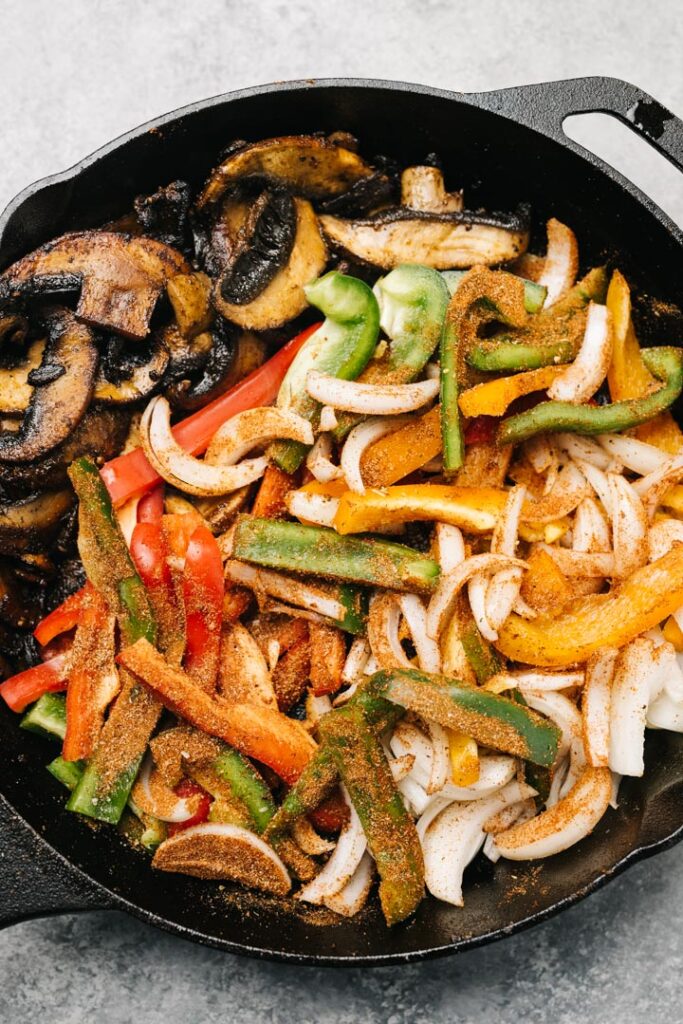 Sliced onions and bell peppers sprinkled with fajita seasoning in one half of a cast iron skillet, with sauteed mushrooms in the other half.