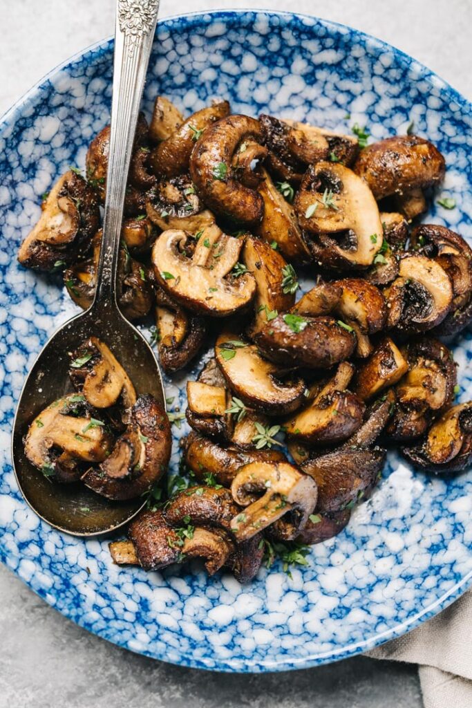 Air fryer mushrooms in a blue speckled bowl with a vintage silver serving spoon, garnished with fresh thyme.