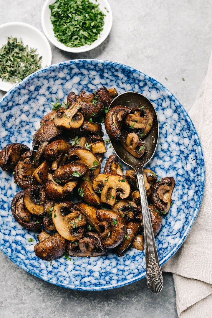 Air fryer mushrooms in a blue speckled bowl on a concrete background, with small bowls of fresh herbs and a tan linen napkin.