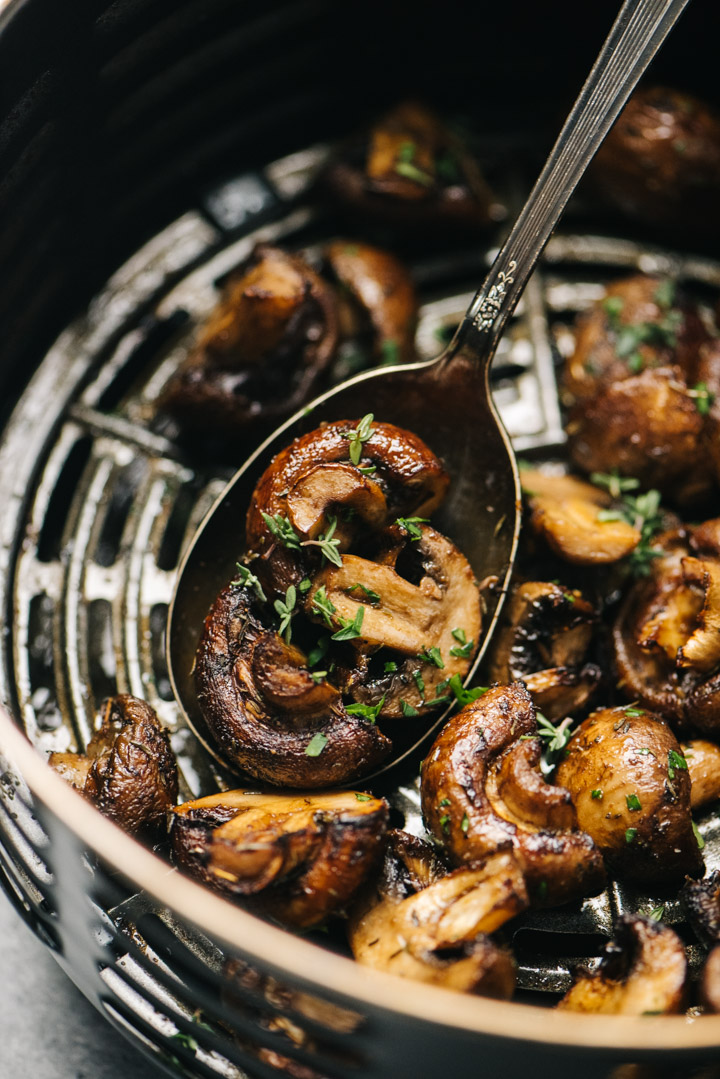 Side view, a silver serving spoon tucked into air fried mushrooms in the basket of an air fryer.