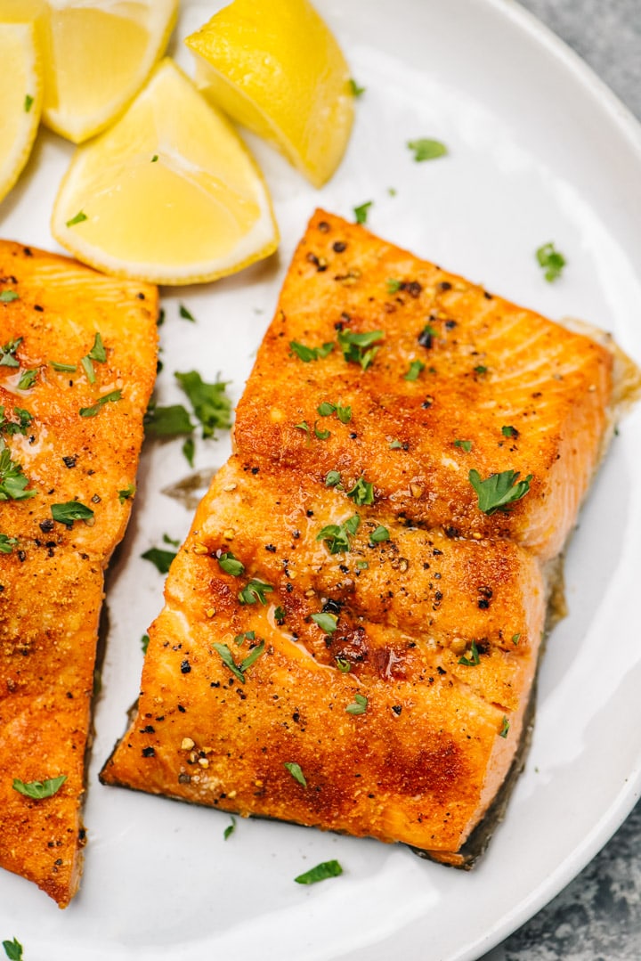 Two pieces of salmon cooked in the air fryer on a white plate, garnished with lemon wedges and fresh chopped parsley.