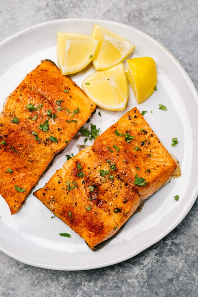 Two pieces of air fryer salmon on a white plate with lemon wedges and chopped parsley.