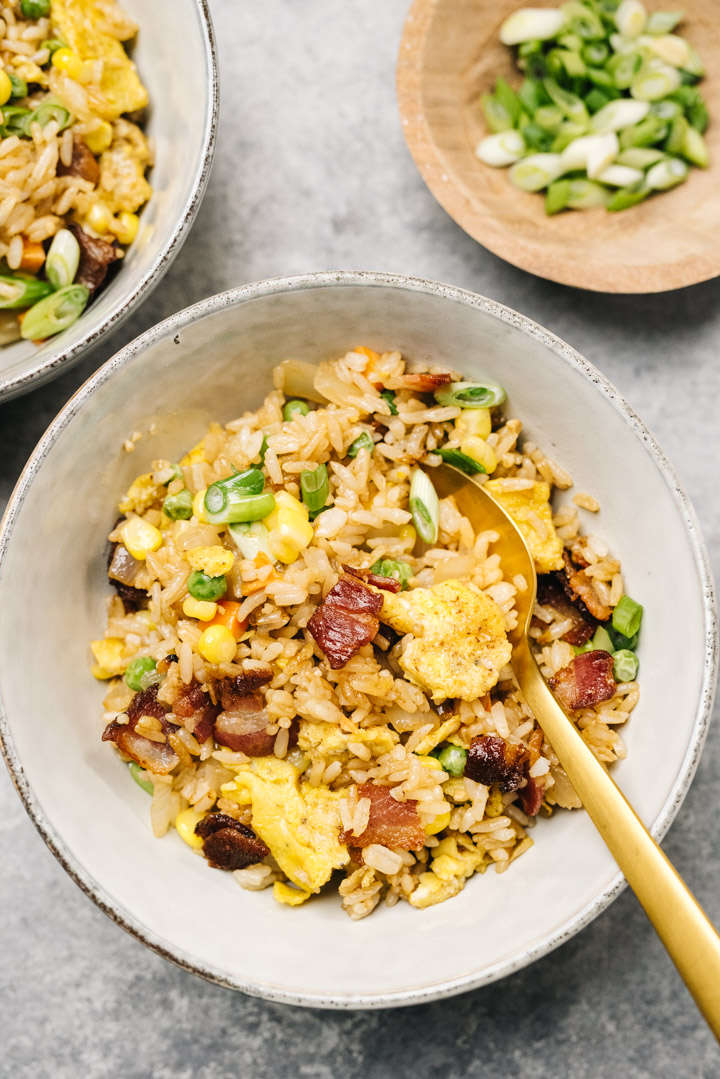 Two bowls of fried rice with bacon on a concrete background with a small bowl of chopped green onions.