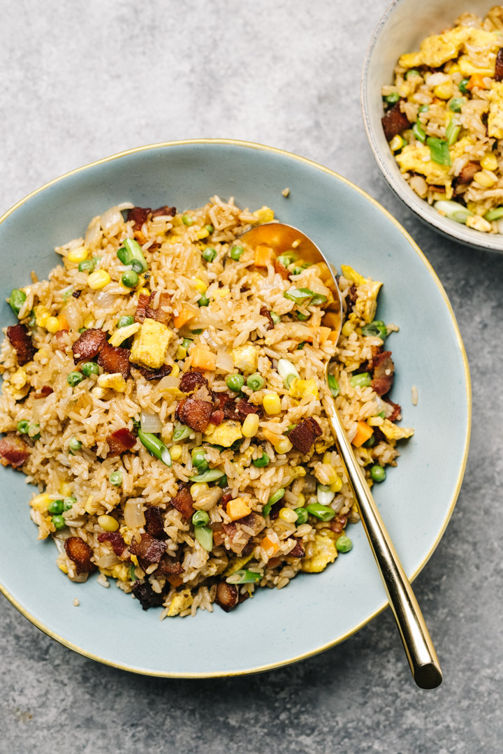A large blue bowl of fried rice with bacon on a concrete background, with a gold serving spoon.