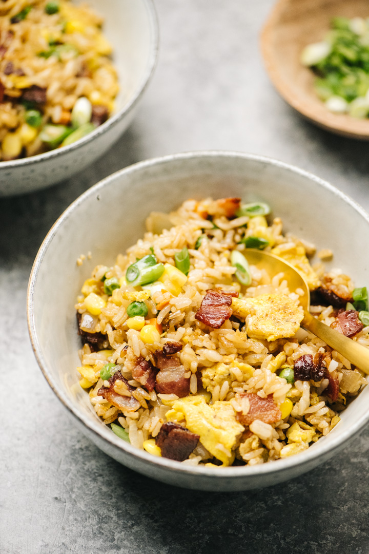 Side view, two bowls of breakfast fried rice on a concrete background with a small wood bowl of chopped green onions in the background.