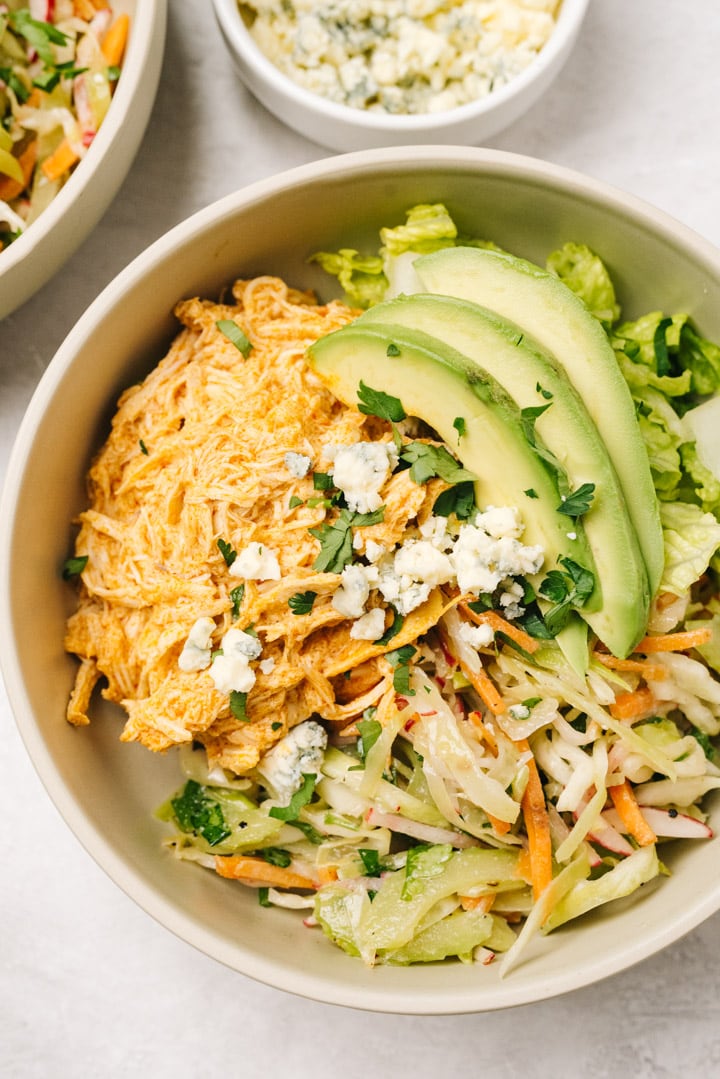 Shredded buffalo chicken with coleslaw and chopped romaine in a tan bowl, topped with avocado slices and blue cheese crumbles.