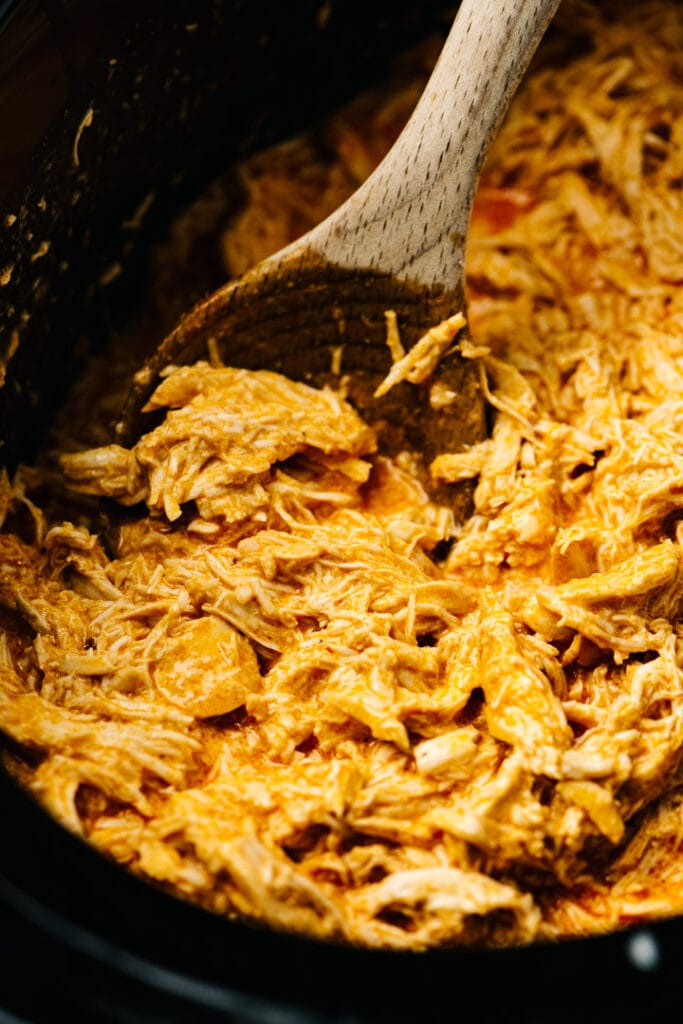 Side view, shredded buffalo chicken in a crockpot with a wood spoon.