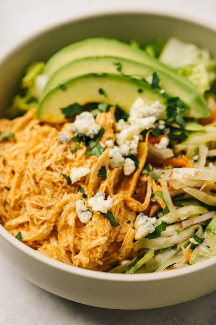 Side view, shredded buffalo chicken over coleslaw and romaine lettuce in a bowl with avocado slices and crumbled blue cheese.