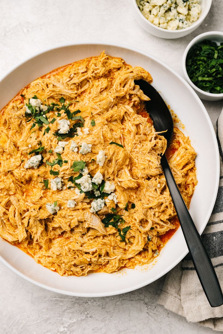 Shredded buffalo chicken in a white serving bowl with a black spoon, with a linen napkin and small bowls of blue cheese and chopped parsley to the side.